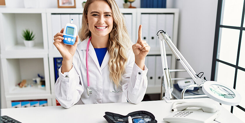Young beautiful doctor woman holding glucose meter smiling with an idea or question pointing finger with happy face, number one