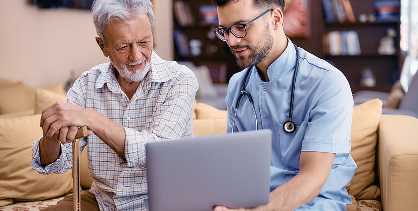 Senior man using laptop with assistance of male caregiver during home visit.